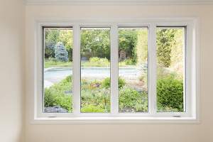 A set of four casement windows with white frame, in a white wall, with a view of a green yard and trees outside.