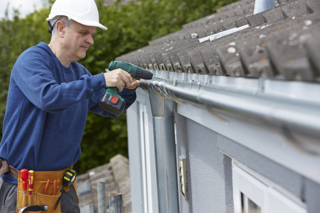 Technician replacing gutters on exterior of a house. Standing on a ladder, using a drill, wearing a white hardhat.